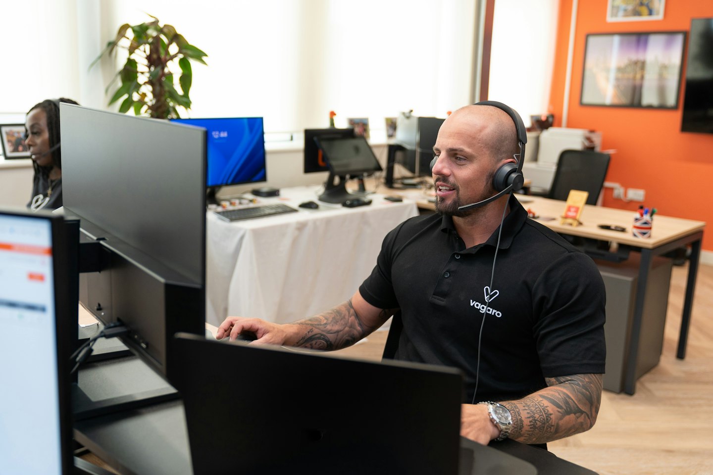 Man with beard and tattoos on headset working at computer monitors.